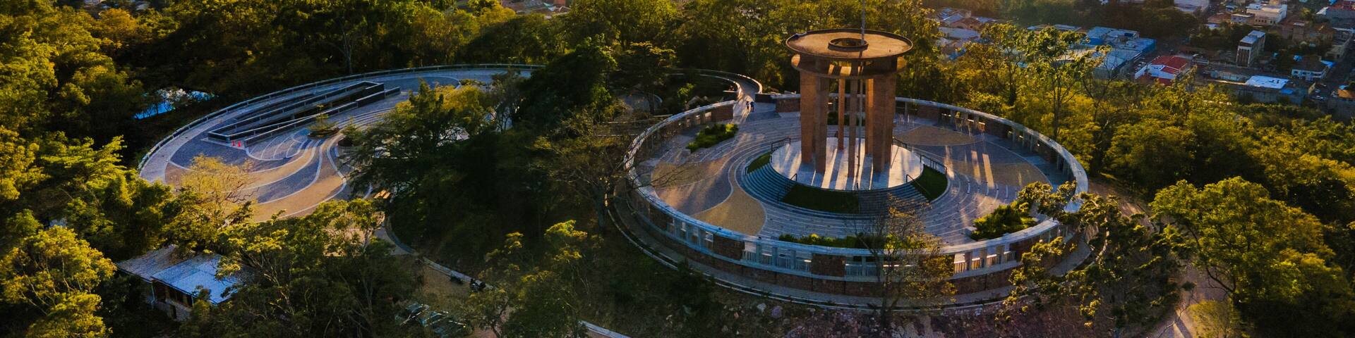 A circular monument with a flag, surrounded by trees and a cityscape in the background.
