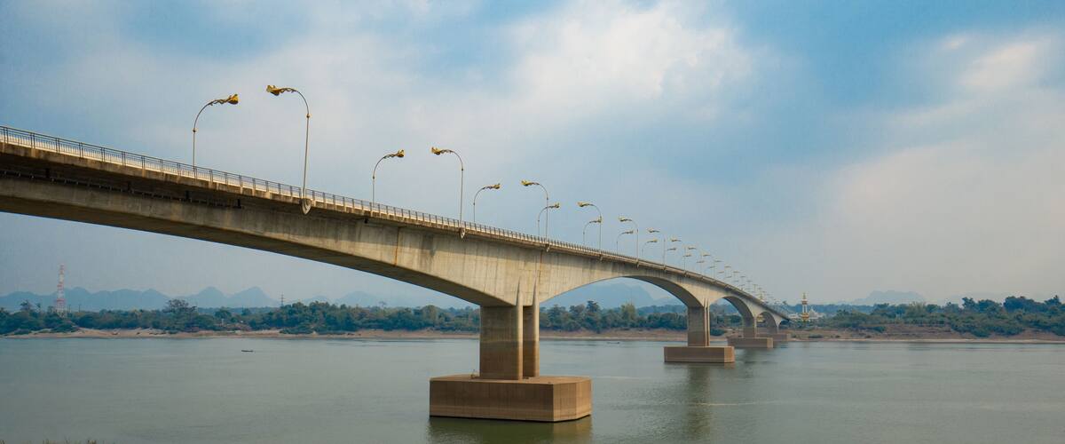 A concrete bridge with multiple arches spanning a river, surrounded by greenery and distant mountains.