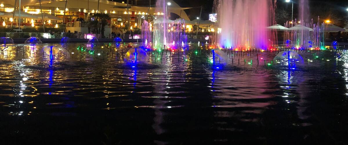 A nighttime fountain with colorful lights reflecting on the water, a dome-shaped building, and a city skyline in the background.