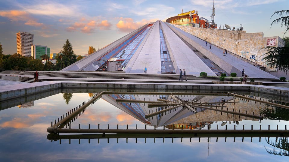 A pyramid-like structure with a reflective pool in front, surrounded by modern buildings and a stadium.