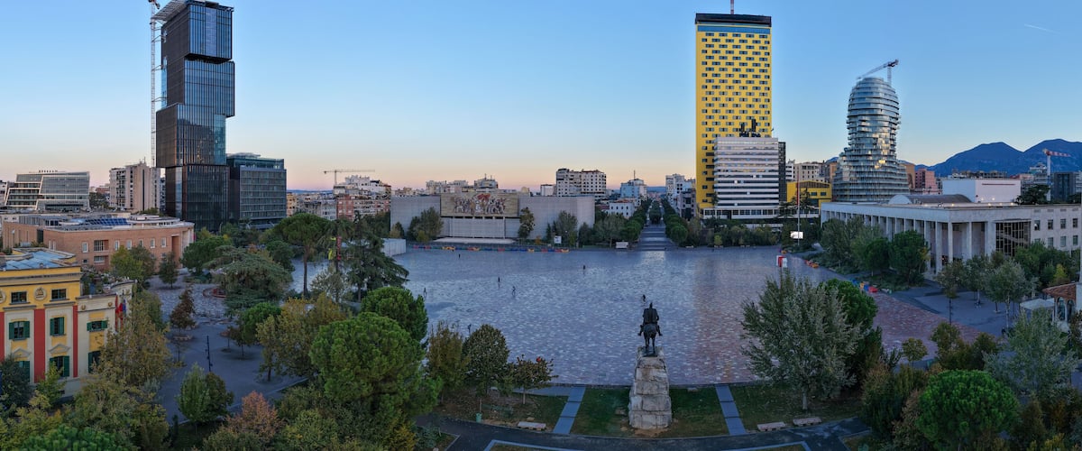 A cityscape with modern buildings, a central square with a statue, and a body of water.