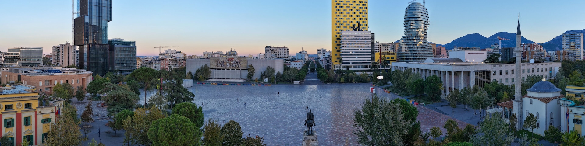 A cityscape with modern buildings, a central square with a statue, and a body of water.