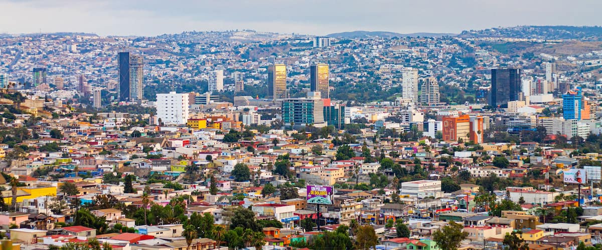 A cityscape with a mix of high-rise buildings and smaller houses, a hilly background, and a cloudy sky.