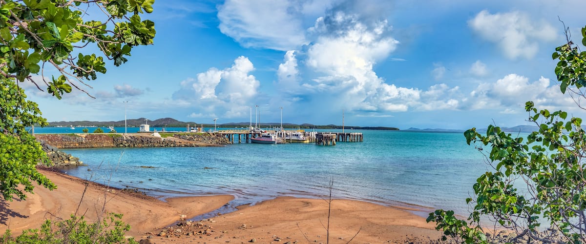 A coastal scene with a sandy beach, calm turquoise waters, a pier with boats, and distant mountains.