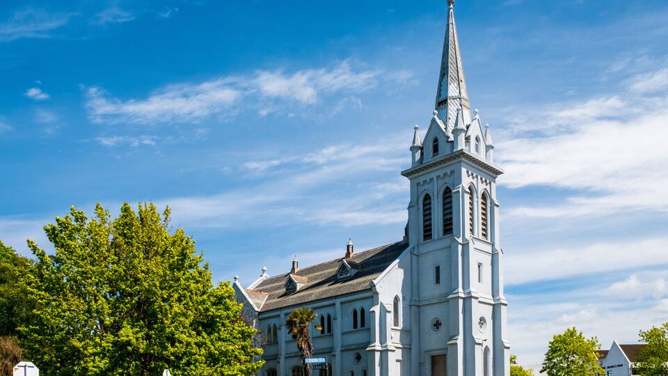 A white church with a tall steeple and a clock tower, surrounded by trees and a clear blue sky.