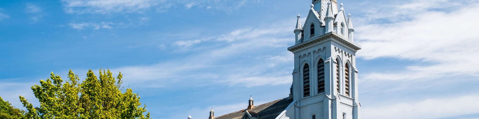 A white church with a tall steeple and a clock tower, surrounded by trees and a clear blue sky.