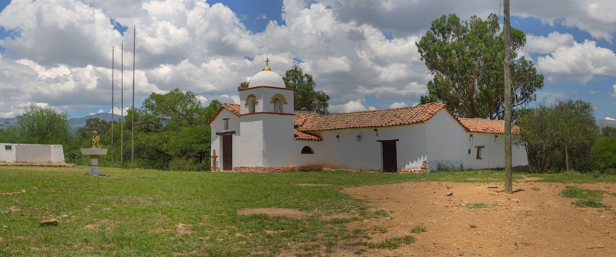 A small white church with a red-tiled roof and a bell tower, surrounded by green grass and trees, with mountains in the background.