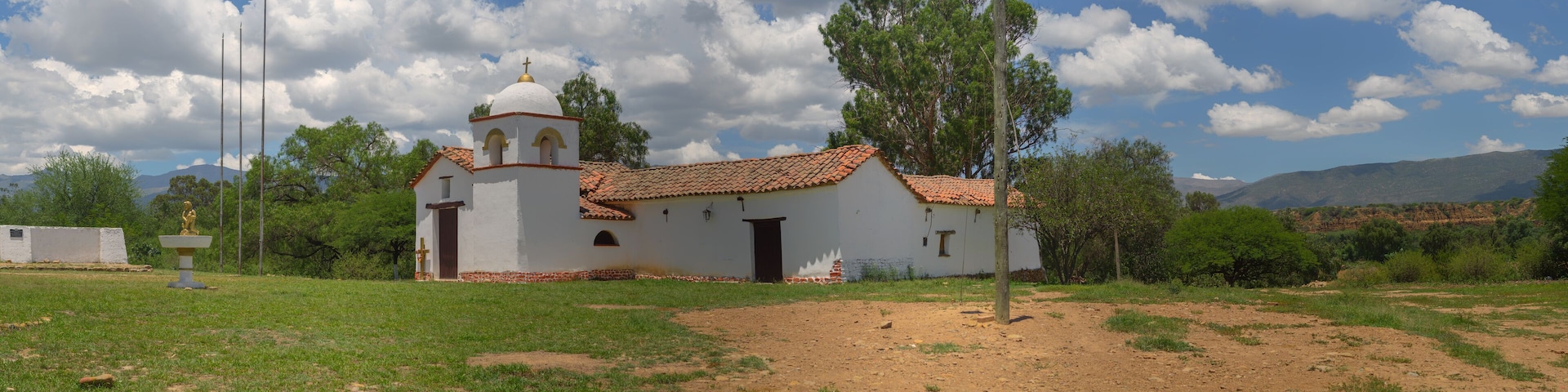 A small white church with a red-tiled roof and a bell tower, surrounded by green grass and trees, with mountains in the background.