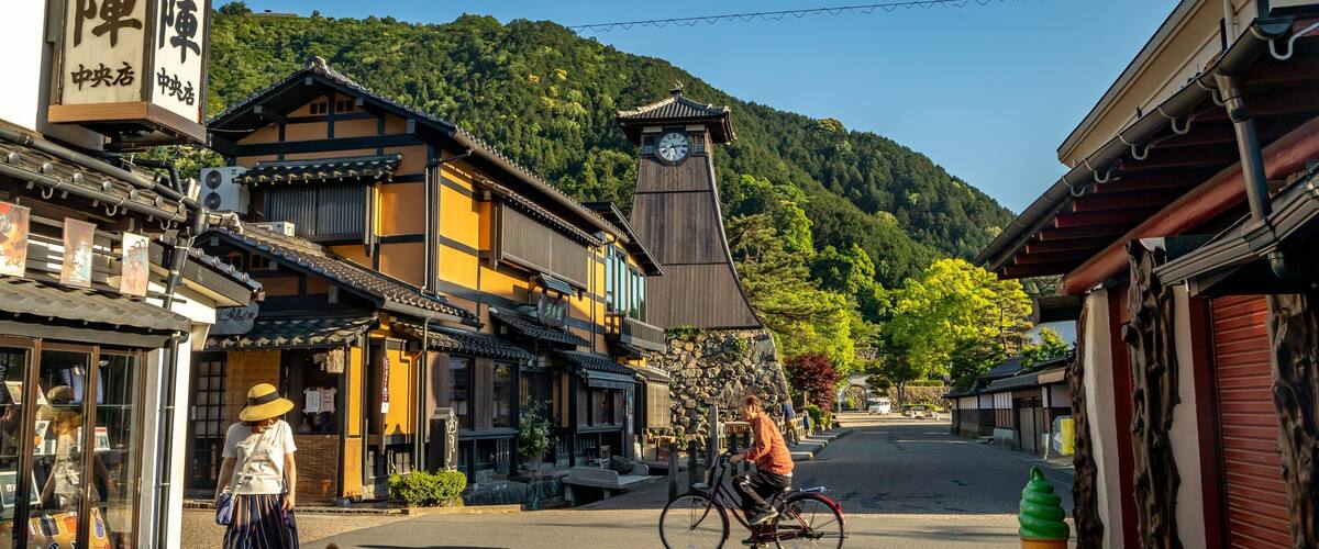 A traditional Japanese street with buildings, a clock tower, and a person riding a bicycle.