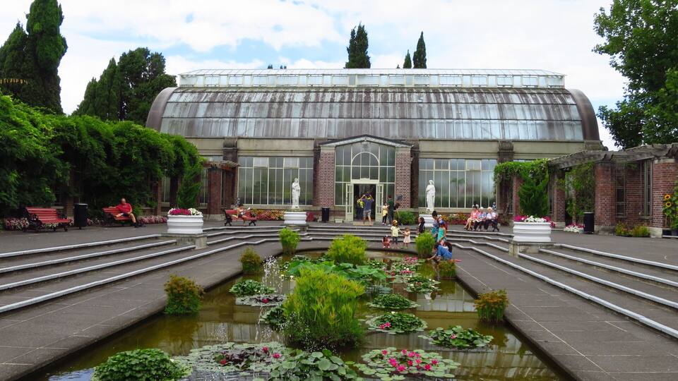 Un invernadero amplio con techo de vidrio, rodeado de un jardín formal con un estanque central y estatuas.