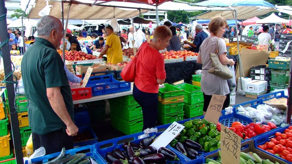 Ein lebhafter Markt mit zahlreichen Ständen, auf denen frisches Obst und Gemüse wie Tomaten, Gurken und Auberginen angeboten werden.