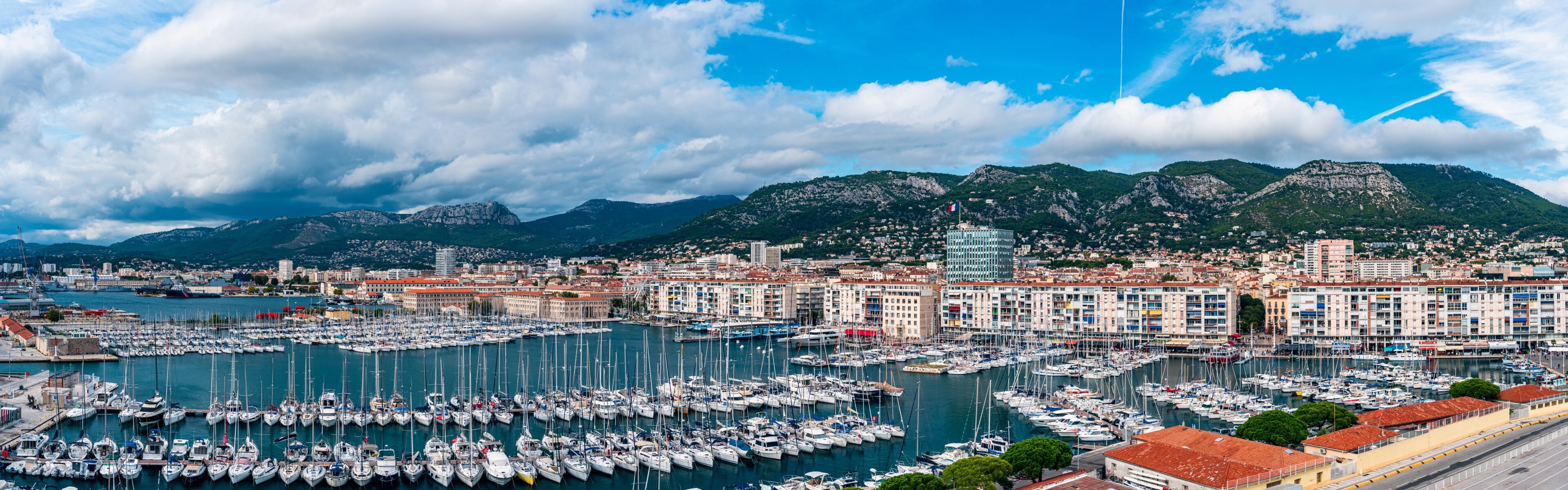 A marina filled with boats, a coastal cityscape with buildings, and mountains in the background.