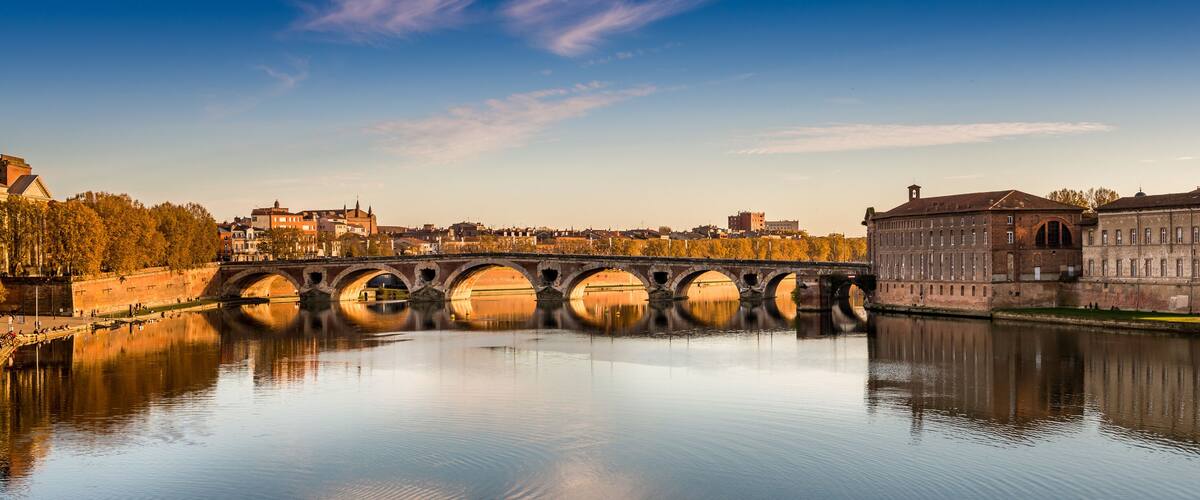 A stone bridge over a calm river, reflecting the surrounding buildings and trees.
