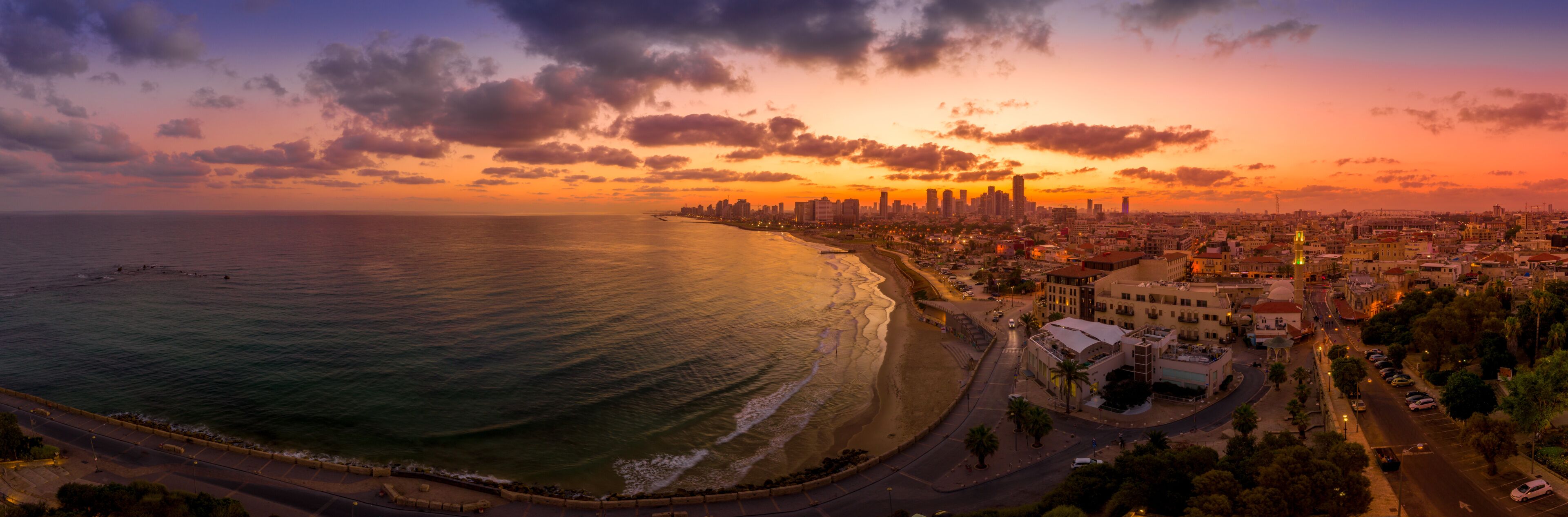 A coastal cityscape at sunset with a prominent skyline, a beach, and a road along the coast.