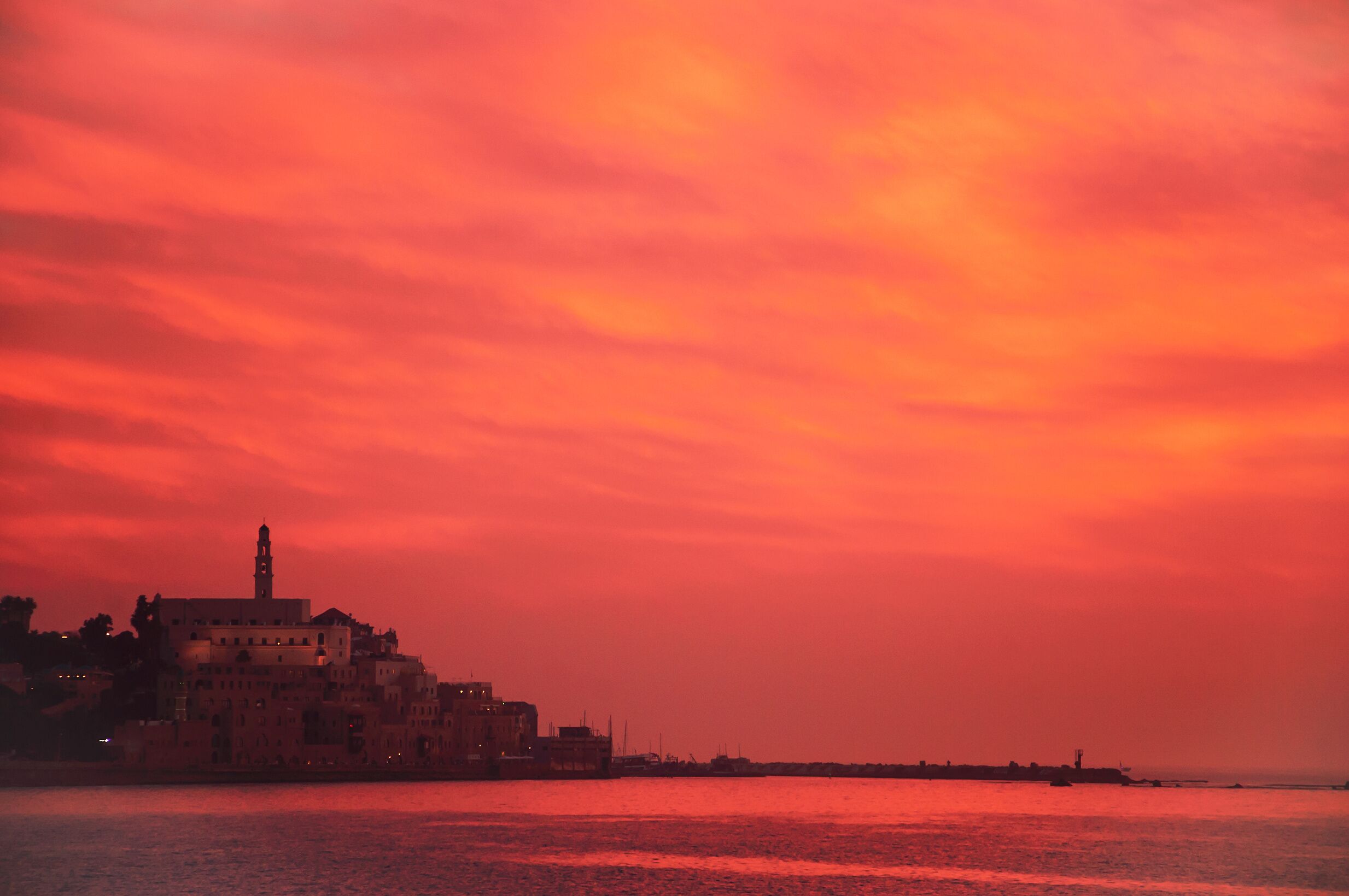 Un paisaje urbano costero al atardecer con una torre destacada y edificios a lo largo del paseo marítimo.