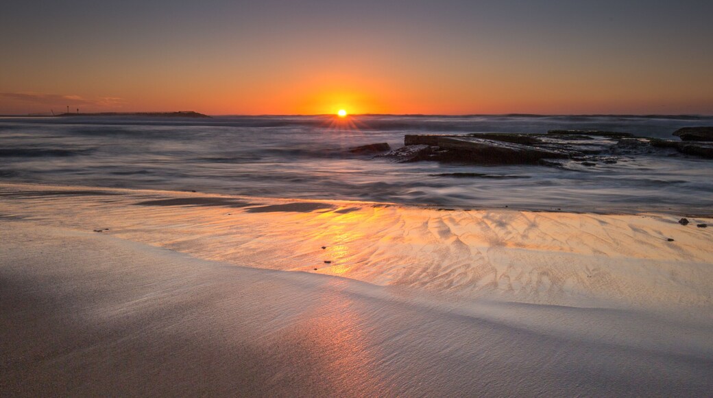 Uma praia ao pôr do sol com ondas, um cais e um farol.