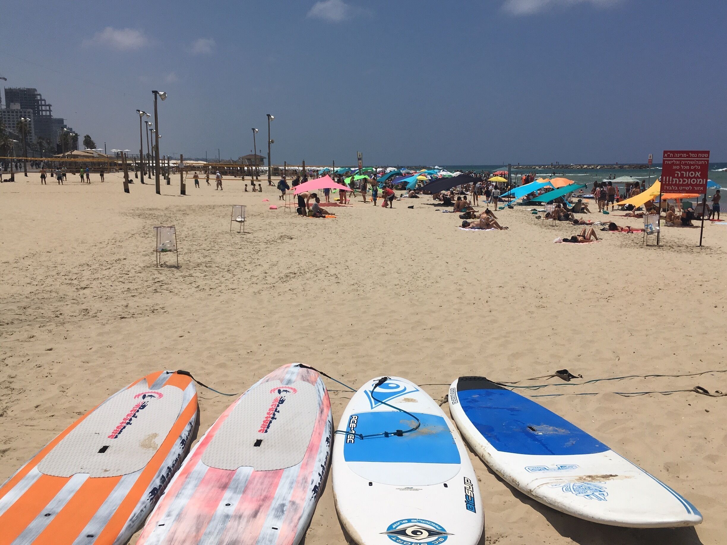 A beach with surfboards lined up in the foreground, umbrellas, and people relaxing on the sand.