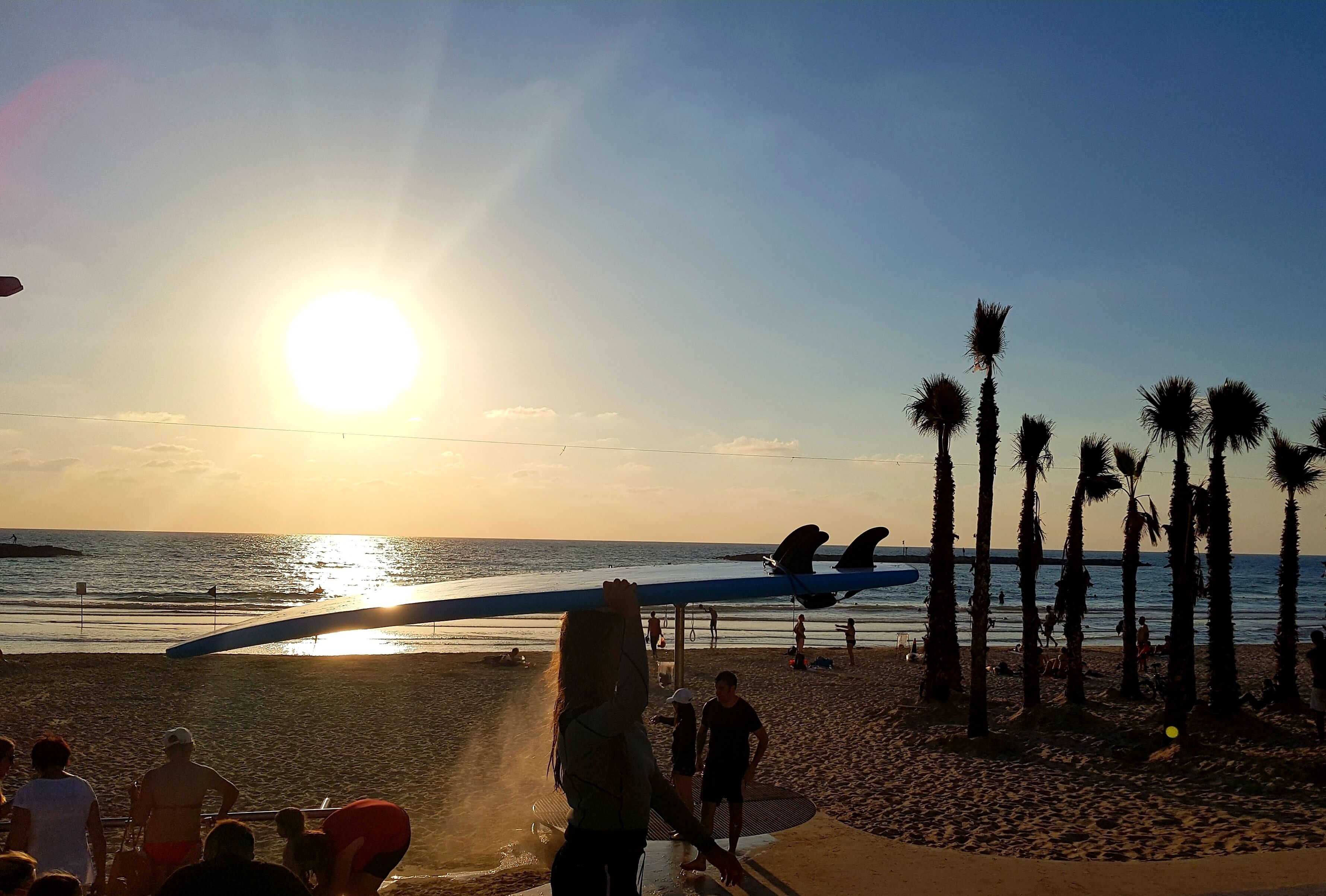 Une plage au coucher du soleil, avec des palmiers, une planche de surf et des gens.