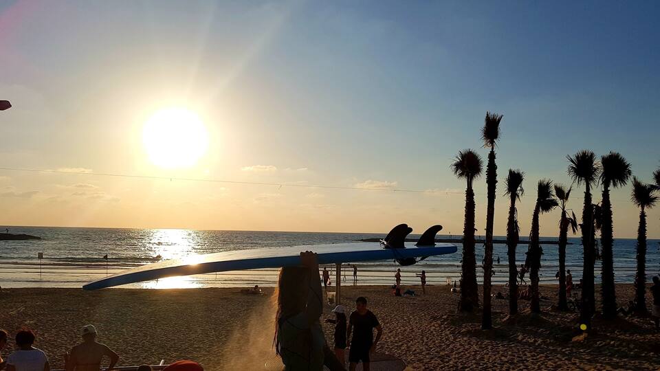 Une plage au coucher du soleil, avec des palmiers, une planche de surf et des gens.