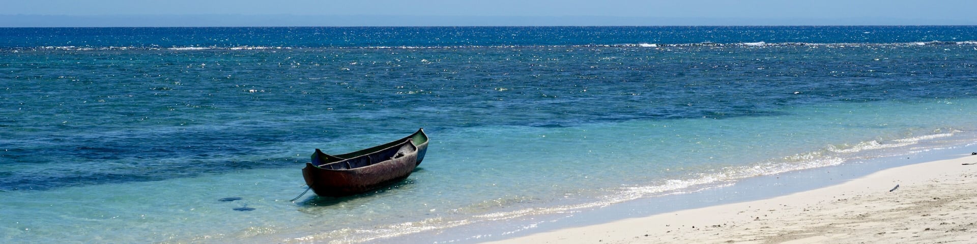 A boat on a sandy beach with clear blue water and a calm sea.