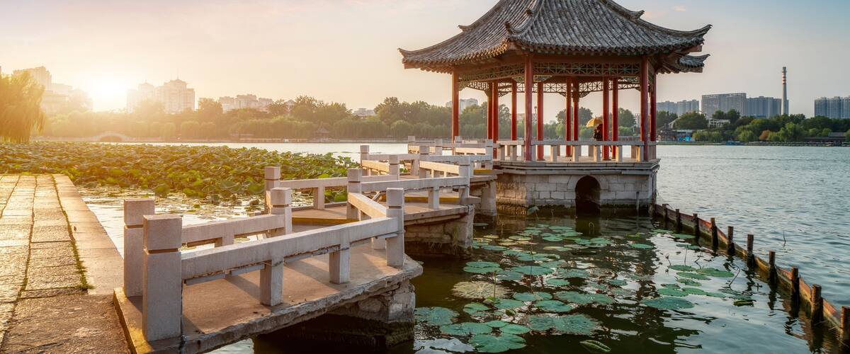 A traditional pavilion by a calm water body with lily pads, a stone bridge, and a city skyline in the background.