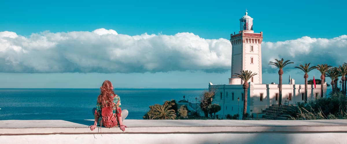 A woman sitting by a coastal road with a lighthouse and palm trees in the background.