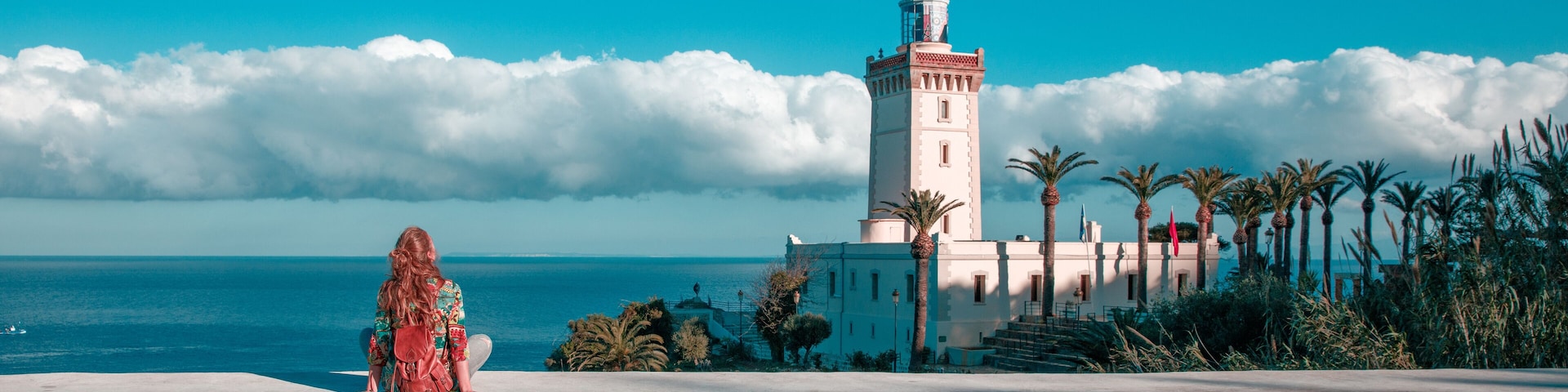 A woman sitting by a coastal road with a lighthouse and palm trees in the background.