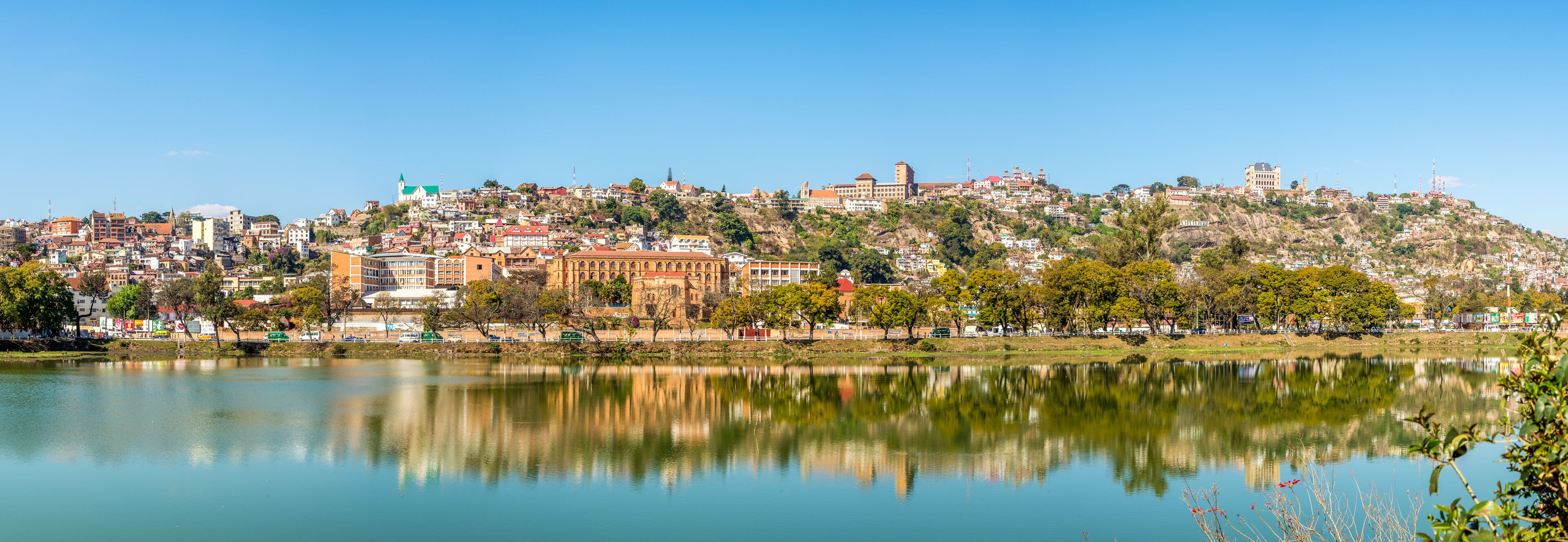 A cityscape with a hilltop castle, a calm lake reflecting the buildings, and a clear blue sky.