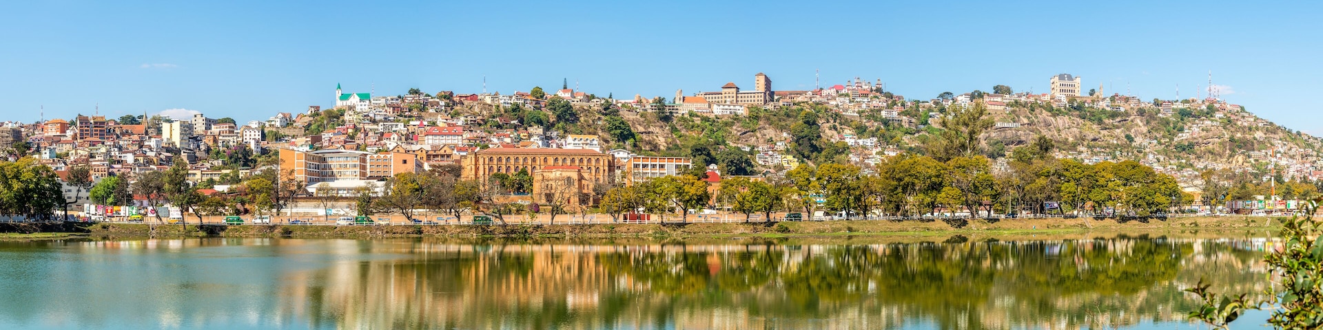 A cityscape with a hilltop castle, a calm lake reflecting the buildings, and a clear blue sky.