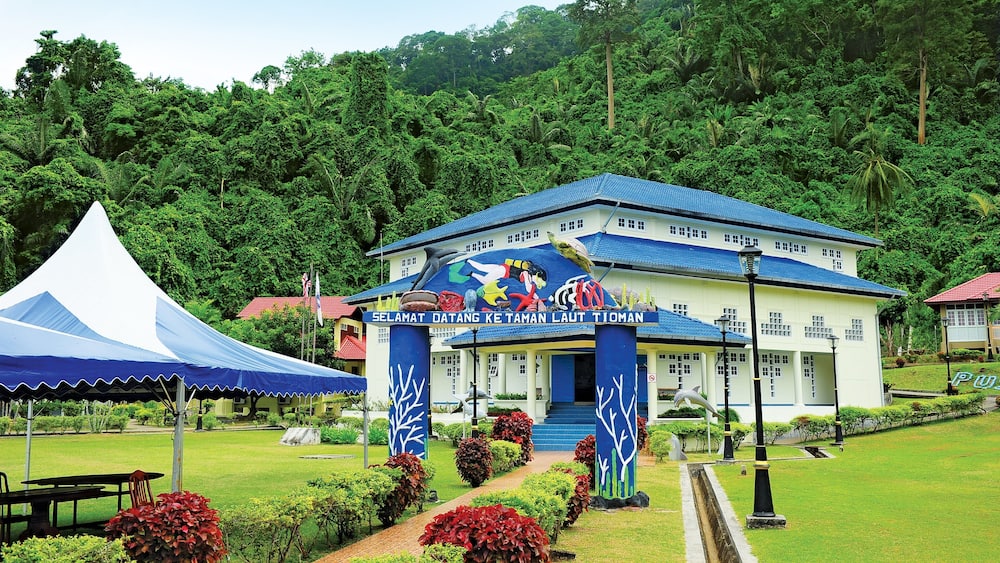 Un bâtiment avec un toit bleu et une fresque murale colorée. On y voit une tente et un jardin avec des arbustes rouges.