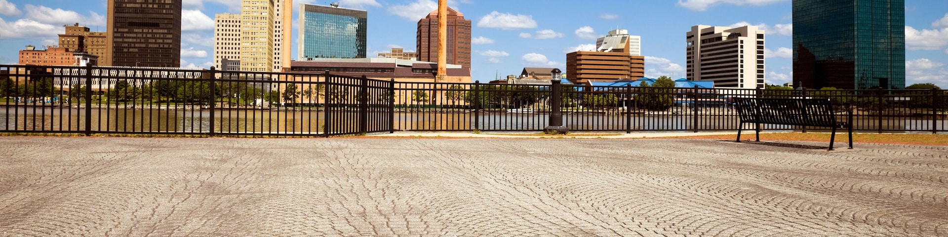 A cityscape with a cobblestone plaza, a black metal fence, and modern buildings under a blue sky with clouds.