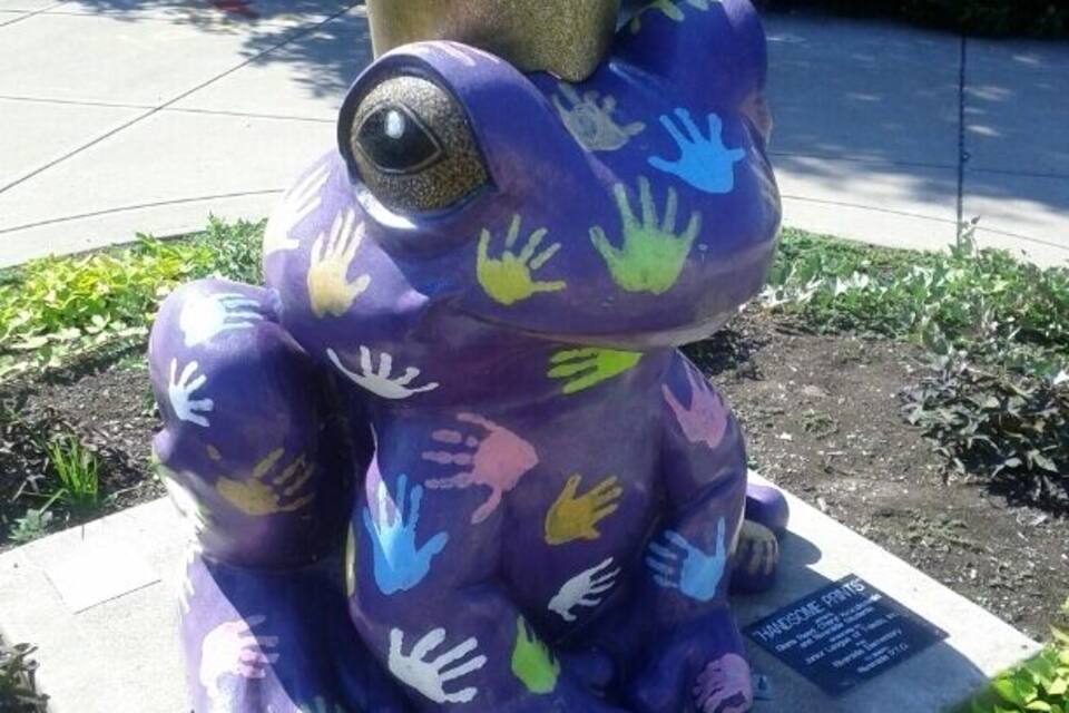 A purple frog statue with a crown and handprints on its body, placed on a pedestal.