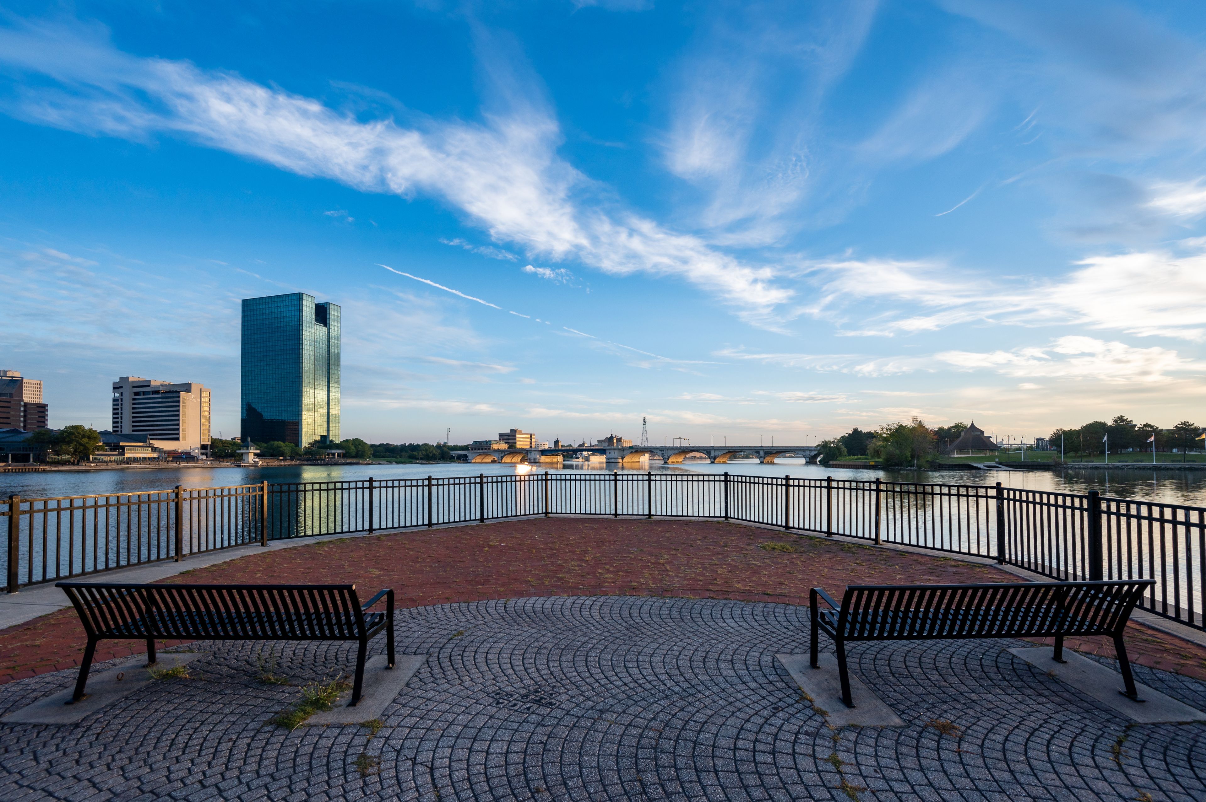 Une promenade le long de la rivière, agrémentée de bancs, un gratte-ciel moderne et un pont en arrière-plan.