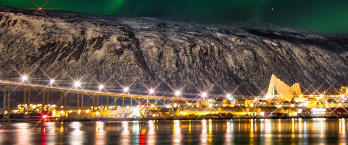The aurora borealis over a coastal city with a bridge and buildings reflecting on the water.