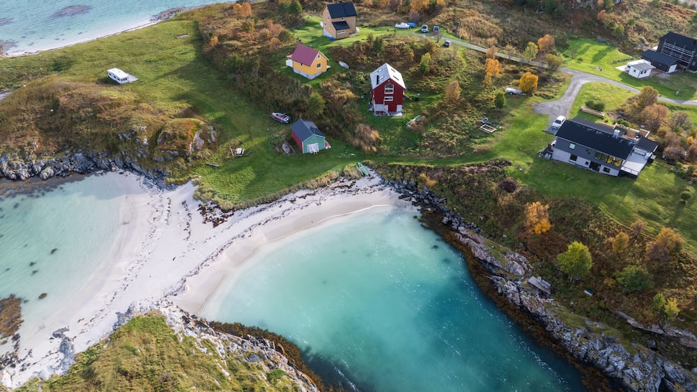 Aerial view of colorful houses scattered on green hills overlooking a white sand beach and turquoise sea at Sommaroy, Troms, Norway.