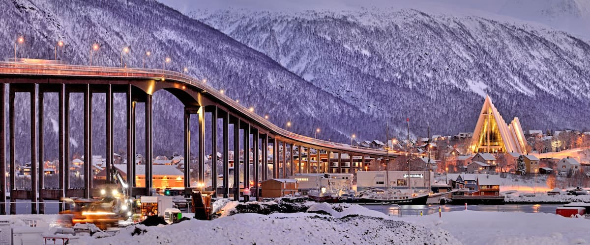Uma ponte coberta de neve com luzes iluminadas, um cenário montanhoso e um edifício moderno com telhado triangular.