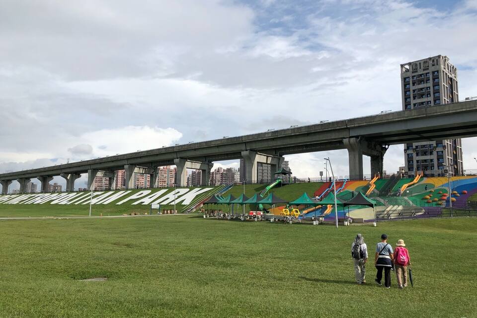 Ein Park mit einer großen, grünen Wiese, einem bunten Spielplatz und einer Brücke im Hintergrund.