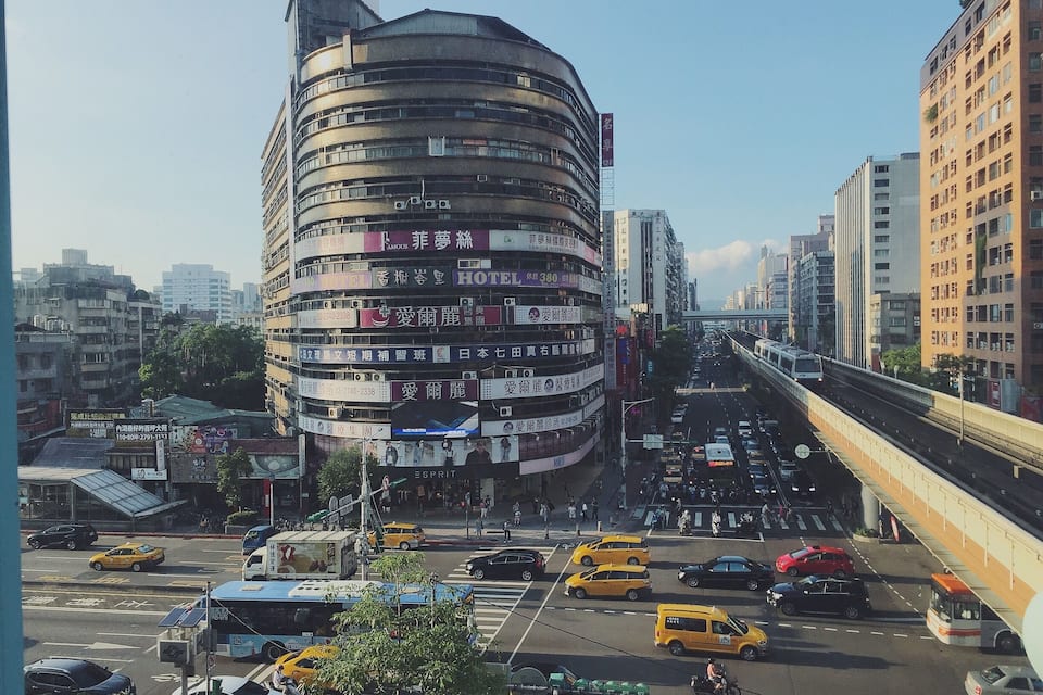 A busy urban street with a distinctive curved building, yellow taxis, and a train on an elevated track.