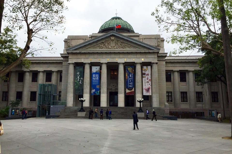A large neoclassical building with a green dome, columns, and banners.