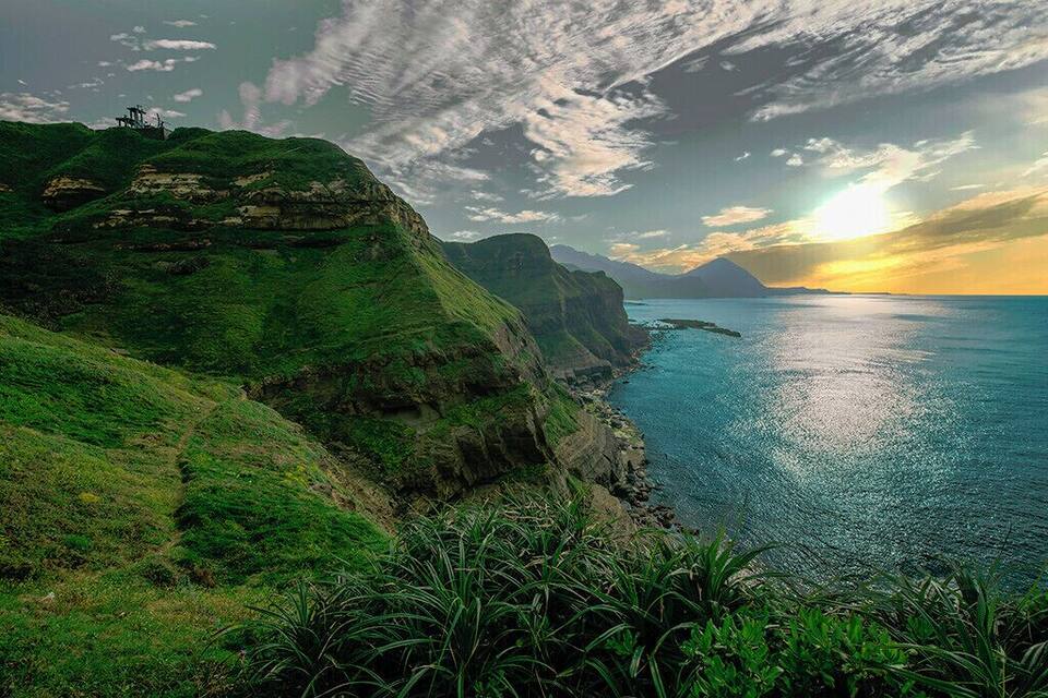 A coastal landscape with green hills, a body of water, and a sunset.