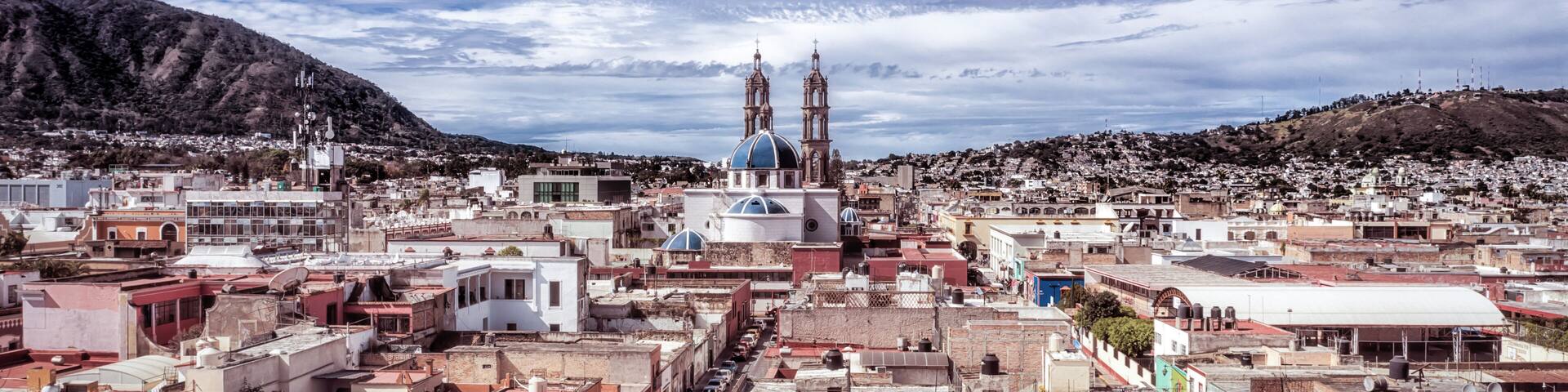 A cityscape with a prominent church with two towers, surrounded by densely packed buildings and a hilly backdrop.