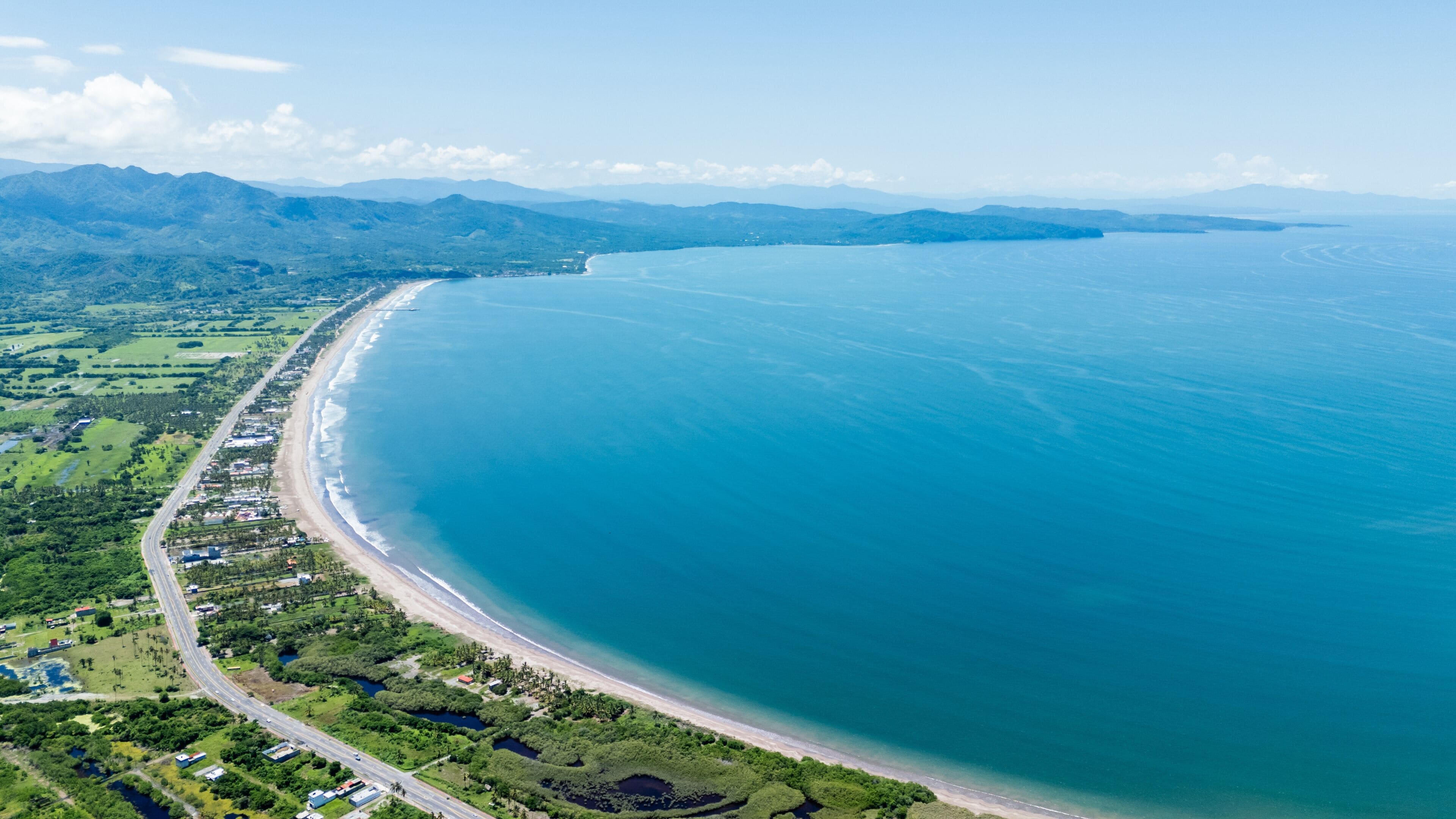Vista aérea de una zona costera con una playa extensa, abundante vegetación y montañas distantes.