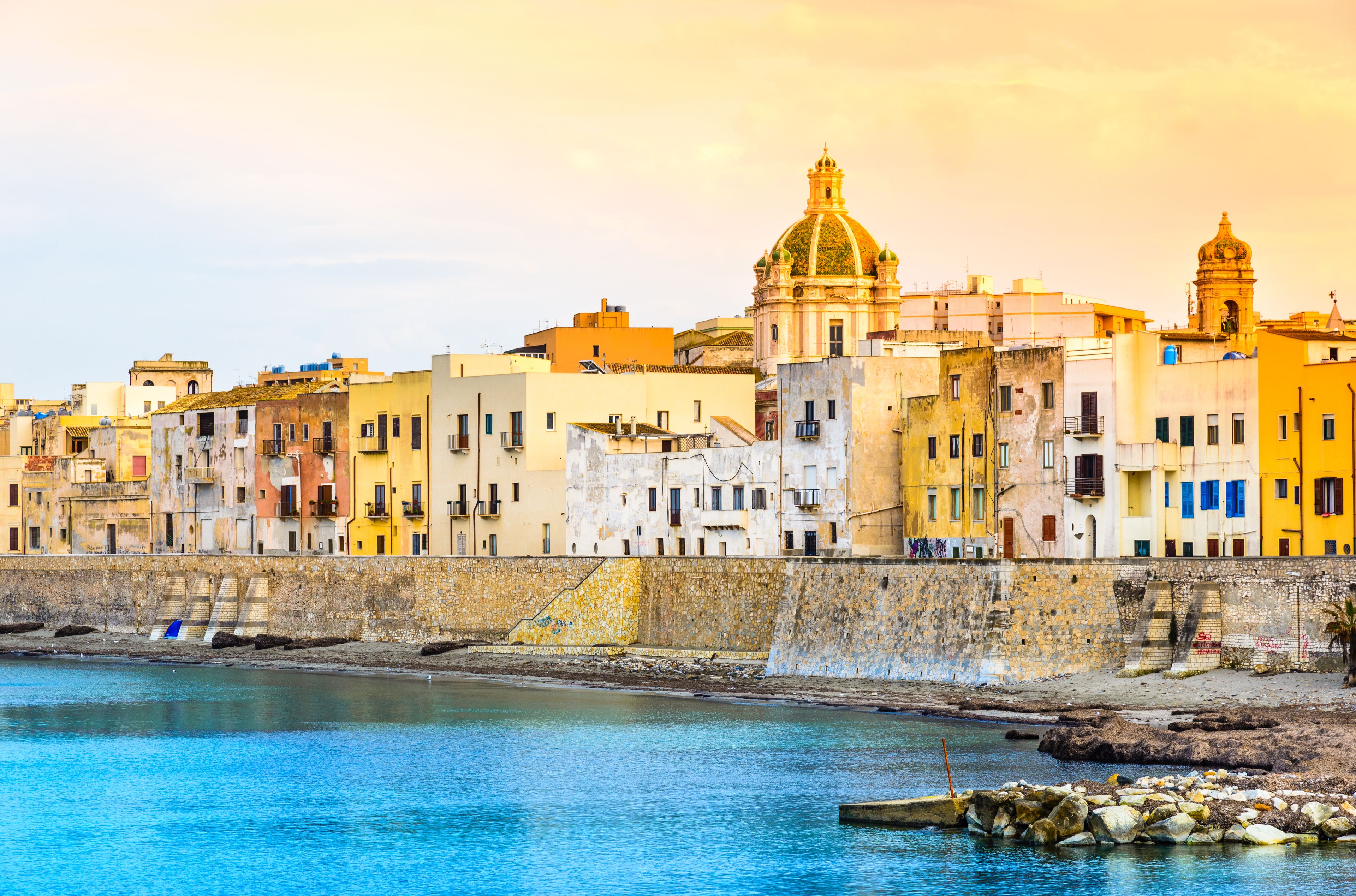 A coastal cityscape with colorful buildings, a prominent church with a golden dome, and a stone wall along the waterfront.