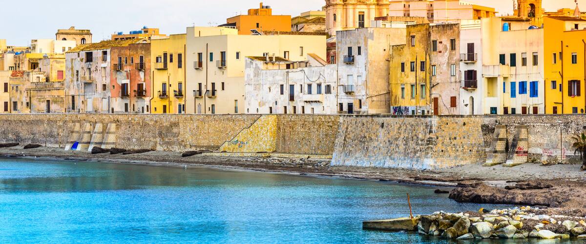A coastal cityscape with colorful buildings, a prominent church with a golden dome, and a stone wall along the waterfront.