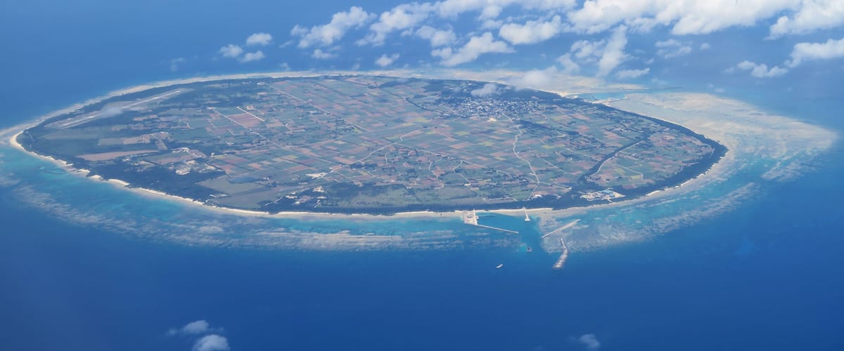 An aerial view of a circular island with a clear coastline, surrounded by ocean and scattered clouds.