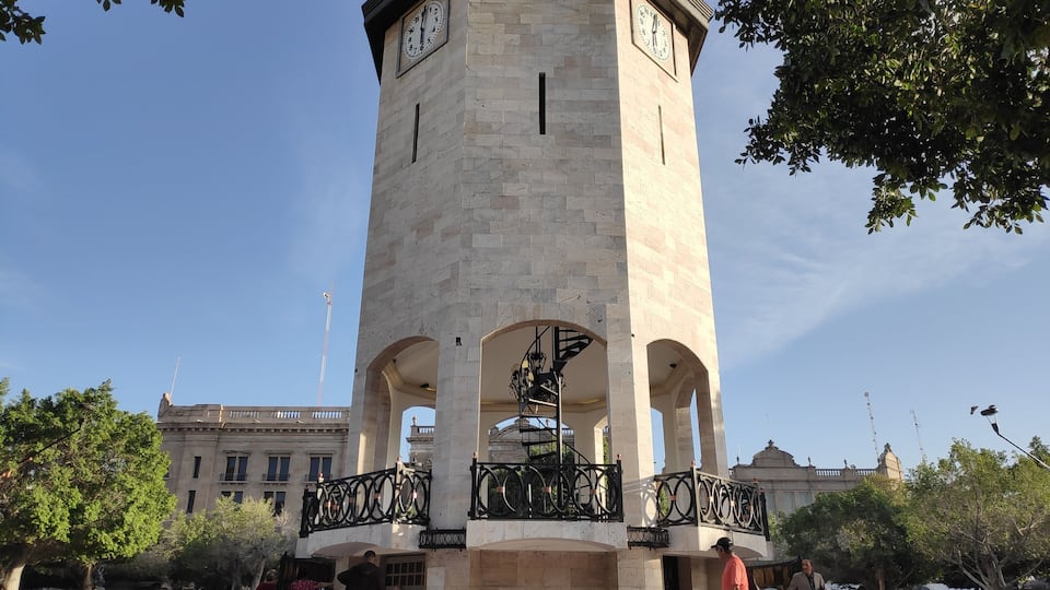 A tall clock tower with a balcony, surrounded by trees and people.