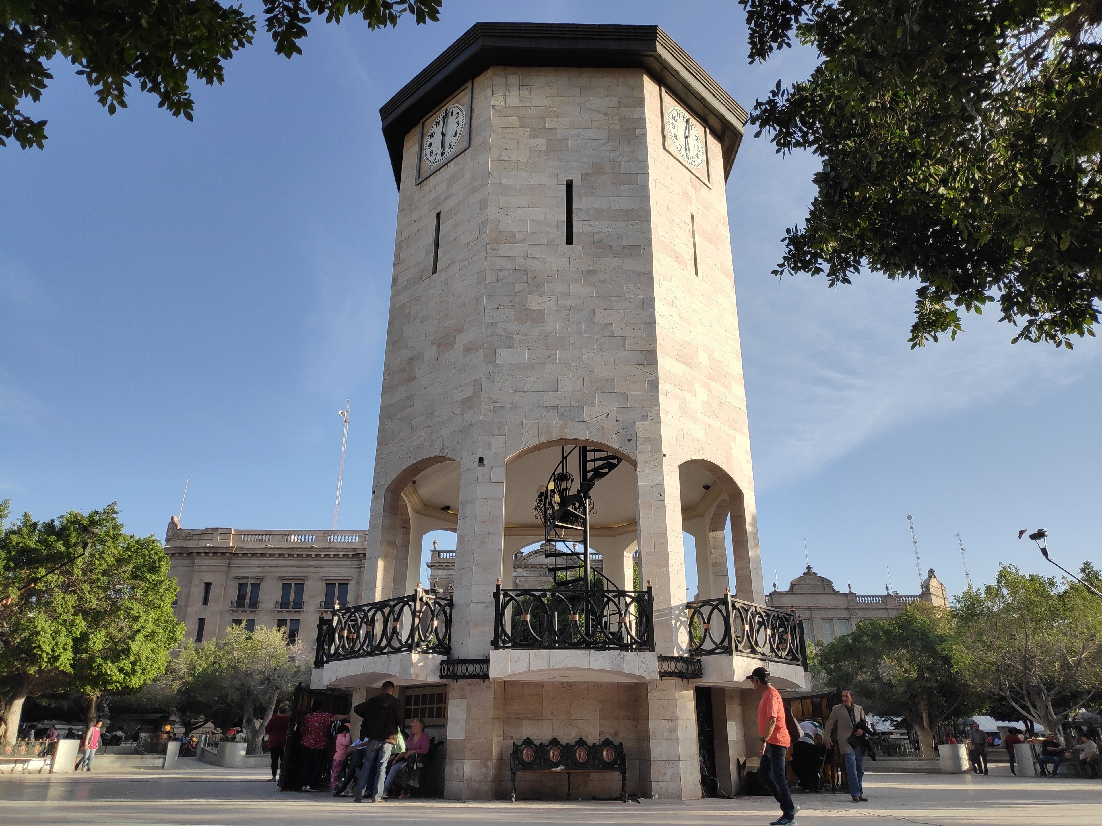 A tall clock tower with a balcony, surrounded by trees and people.