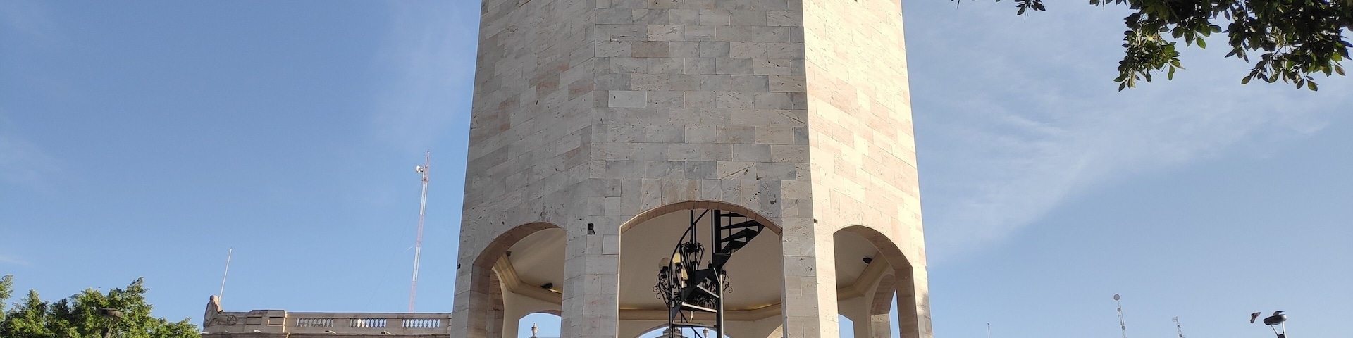 A tall clock tower with a balcony, surrounded by trees and people.