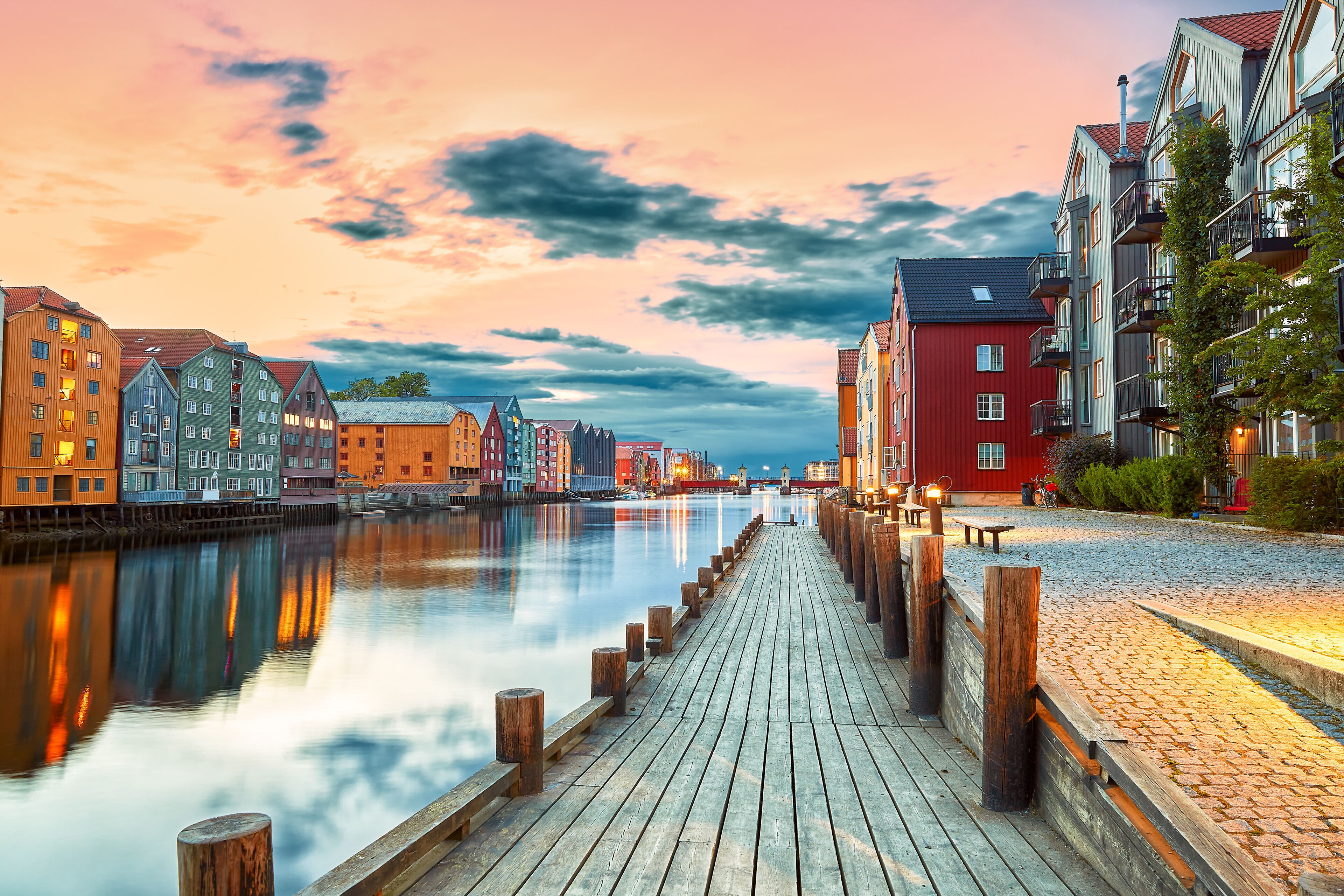 Un paseo marítimo con cubierta de madera, edificios coloridos y un río tranquilo que refleja el cielo.