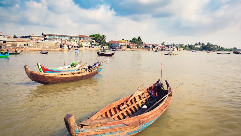 A river with traditional wooden boats, a distant shoreline with buildings, and a partly cloudy sky.